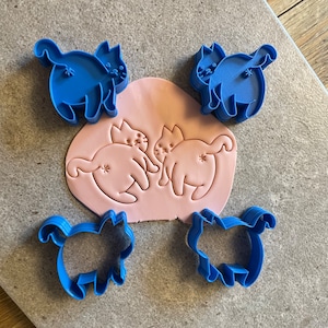 May include: Four blue plastic cat-shaped cookie cutters, two of which are used to create an impression of two cats on a pink surface.