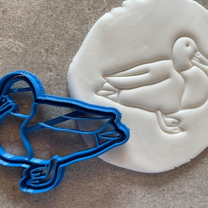 Könnte beinhalten: Ein blauer Plastik-Ausstecher in Form einer Ente. Der Ausstecher liegt auf einer weißen Fondant-Oberfläche mit einer Abdruck einer Ente.