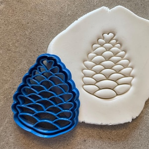 Op de afbeelding: Een blauwe plastic koekjesvormer in de vorm van een dennenappel, naast een witte klei-indruk van de vormer.