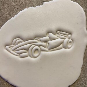 Puede incluir: Un cortador de galletas blanco, de forma ovalada, con un dise&ntilde;o detallado de un coche de carreras de F&oacute;rmula 1. El coche presenta ruedas visibles, una cabina y un aler&oacute;n trasero. El cortador est&aacute; presionado en una masa blanca, creando un relieve en relieve del coche.