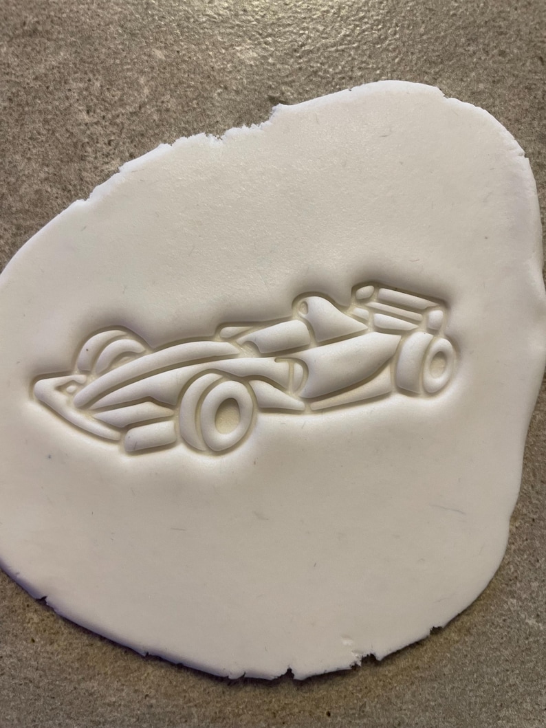 Puede incluir: Un cortador de galletas blanco, de forma ovalada, con un dise&ntilde;o detallado de un coche de carreras de F&oacute;rmula 1. El coche presenta ruedas visibles, una cabina y un aler&oacute;n trasero. El cortador est&aacute; presionado en una masa blanca, creando un relieve en relieve del coche.