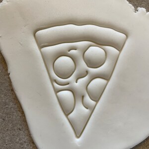 May include: A white clay surface with an embossed image of a pizza slice. The pizza slice has a crust, cheese, and pepperoni.