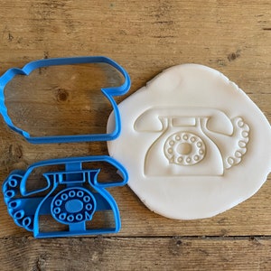 May include: A blue plastic cookie cutter and a dough impression, both in the shape of a vintage rotary phone. The cutter has a detailed design of the phone's dial and handset. The items are on a wooden surface.