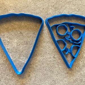 May include: Two blue plastic cookie cutters shaped like pizza slices. One cutter is plain and the other has a design of pepperoni slices.