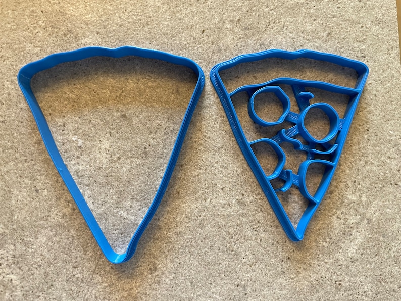 May include: Two blue plastic cookie cutters shaped like pizza slices. One cutter is plain and the other has a design of pepperoni slices.