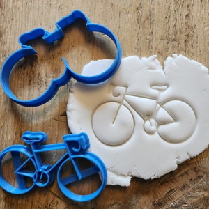 May include: Blue plastic cookie cutters in the shape of a bicycle. One cutter is used to create the outline of a bicycle, and the other cutter is used to create the details of the bicycle, such as the wheels and handlebars. The cutters are shown on a white surface.