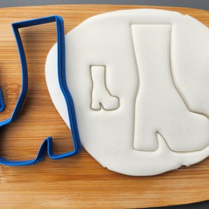 May include: A blue plastic boot-shaped cookie cutter next to a smaller version and a white dough cutout of a boot. The items are on a wooden cutting board, with a gray background.