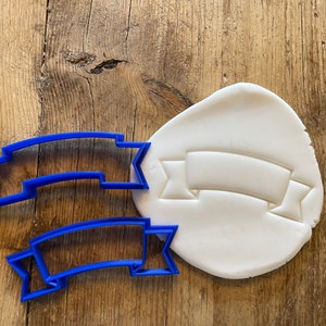 May include: Two blue plastic cookie cutters shaped like a banner with pointed ends. The cutters are on a wooden surface with a white piece of modeling clay that has been pressed with the banner cutter.