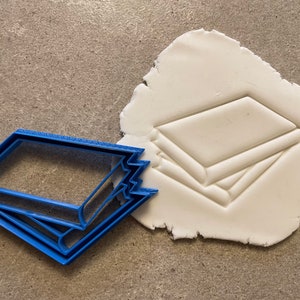 Könnte beinhalten: Ein blauer Plastik-Ausstecher in Form eines Stapels von drei Büchern. Der Ausstecher ist neben einem weißen Abdruck des Ausstechers in einem Stück Modelliermasse zu sehen.