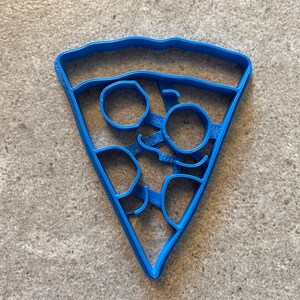 May include: A blue plastic cookie cutter shaped like a slice of pizza with pepperoni and cheese.