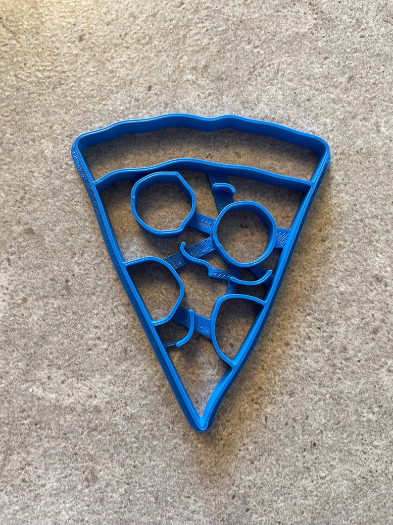May include: A blue plastic cookie cutter shaped like a slice of pizza with pepperoni and cheese.