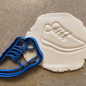 May include: Blue plastic cookie cutter shaped like a trainer. The cutter is shown next to a white impression of the trainer in a piece of modelling clay.
