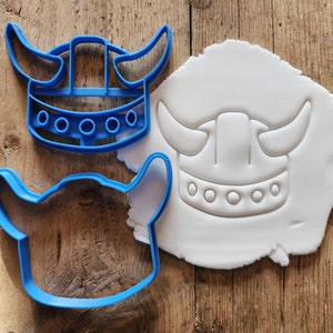Könnte beinhalten: Zwei blaue Plastik-Ausstecher in Form eines Wikingerhelms. Ein Ausstecher ist in weißen Fondant gedrückt.