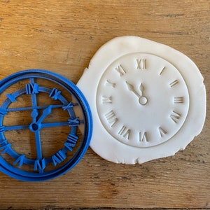 May include: A blue plastic cookie cutter shaped like a clock face with Roman numerals.  A white fondant clock face with Roman numerals is also shown.
