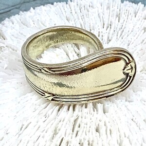 May include: A gold-toned spoon ring with a curved design. The ring is made from a vintage spoon and has a smooth, polished finish.