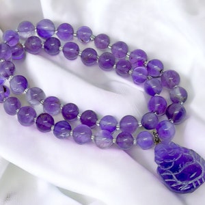 May include: A purple amethyst gemstone necklace with a carved amethyst pendant. The necklace is made of round beads and the pendant is shaped like a Buddha.