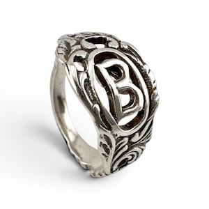 Peut inclure: Une bague en métal argenté présentant un design ajouré avec une lettre majuscule B proéminente au centre. L'anneau est orné de motifs complexes en relief rappelant des volutes et des feuilles qui entourent le motif central de la lettre.