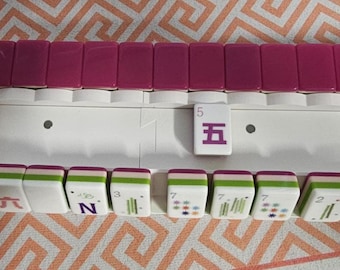 May include: A white plastic Mahjong rack holding a row of magenta tiles along the top edge and a collection of white tiles with various green, blue, and red symbols, numbers, and floral designs arranged in a horizontal line along the bottom shelf.