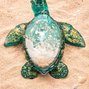 May include: A teal-coloured sea turtle ornament with a transparent shell and green, textured flippers and head. The turtle is on sand, with small seashells scattered around. A decorative item.