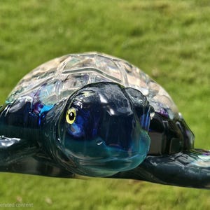 May include: A glass sea turtle figurine featuring a deep blue head and flippers with a translucent, textured shell. The turtle has a prominent yellow eye with a black pupil, and the body displays a glossy finish with subtle iridescent highlights.