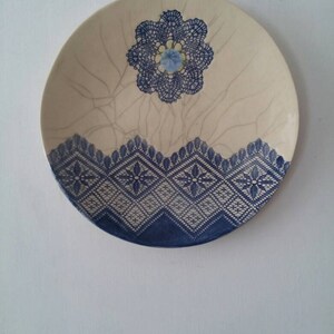 K&ouml;nnte beinhalten: Ein wei&szlig;er Keramikteller mit einem blauen und wei&szlig;en Spitzenmuster. Der Teller hat eine craquel&eacute;e Glasur und eine blaue Blume in der Mitte.