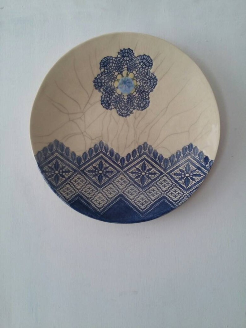 K&ouml;nnte beinhalten: Ein wei&szlig;er Keramikteller mit einem blauen und wei&szlig;en Spitzenmuster. Der Teller hat eine craquel&eacute;e Glasur und eine blaue Blume in der Mitte.