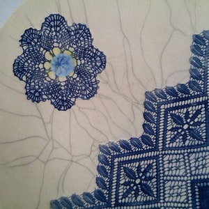 K&ouml;nnte beinhalten: Ein wei&szlig;er Keramikteller mit einem blauen und wei&szlig;en Spitzenmuster. Die Spitze ist in Form einer Blume und eines Quadrats. Der Teller hat eine craquel&eacute;e Glasur.