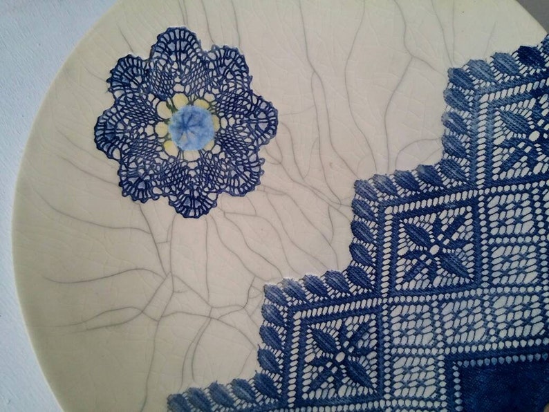 K&ouml;nnte beinhalten: Ein wei&szlig;er Keramikteller mit einem blauen und wei&szlig;en Spitzenmuster. Die Spitze ist in Form einer Blume und eines Quadrats. Der Teller hat eine craquel&eacute;e Glasur.