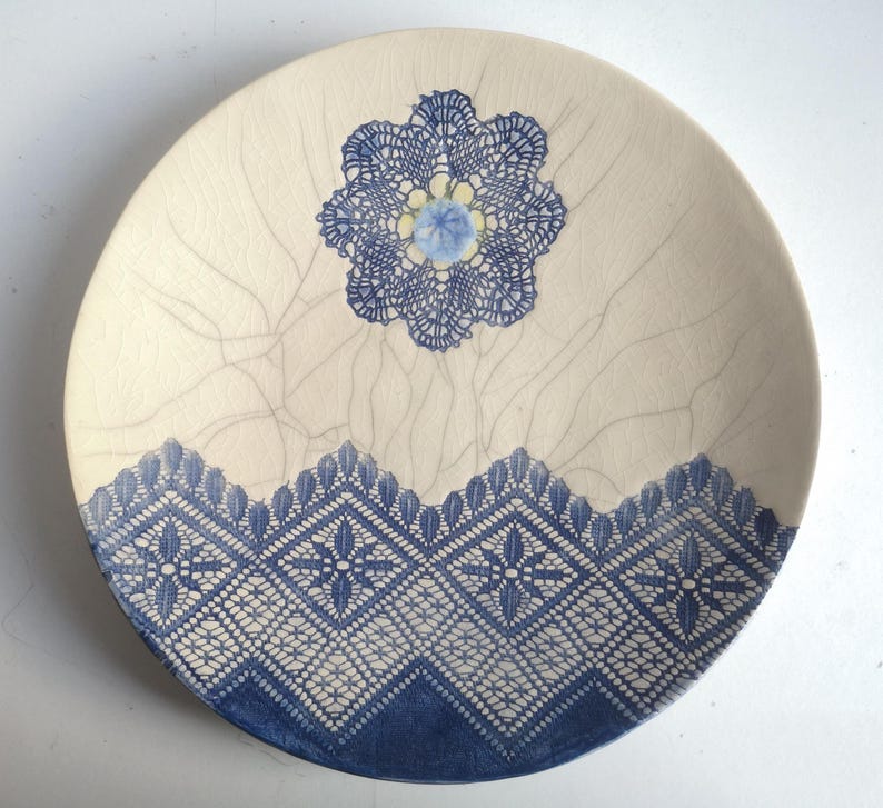 K&ouml;nnte beinhalten: Ein wei&szlig;er Keramikteller mit einem blauen Spitzenmuster. Die Spitze ist in Form einer Blume in der Mitte und eines Randes um den Rand des Tellers.