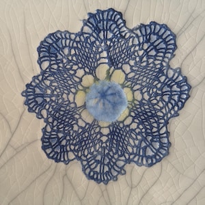 K&ouml;nnte beinhalten: Eine blaue und wei&szlig;e H&auml;keldeckchen mit einem blauen und gelben Blumendesign in der Mitte. Das Deckchen befindet sich auf einem wei&szlig;en Hintergrund mit einer rissigen Textur.