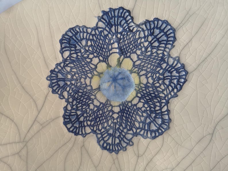 K&ouml;nnte beinhalten: Eine blaue und wei&szlig;e H&auml;keldeckchen mit einem blauen und gelben Blumendesign in der Mitte. Das Deckchen befindet sich auf einem wei&szlig;en Hintergrund mit einer rissigen Textur.