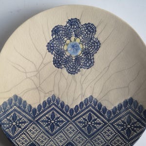 K&ouml;nnte beinhalten: Ein wei&szlig;er Keramikteller mit einem blauen und wei&szlig;en Spitzenmuster. Die Spitze ist in Form einer Blume in der Mitte des Tellers und eines Randes um den Rand.