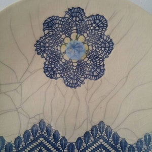 K&ouml;nnte beinhalten: Ein wei&szlig;er Keramikteller mit einem blauen Spitzenmuster. Die Spitze hat die Form einer Blume mit einer blauen Mitte. Der Teller hat eine craquel&eacute;e Glasur.
