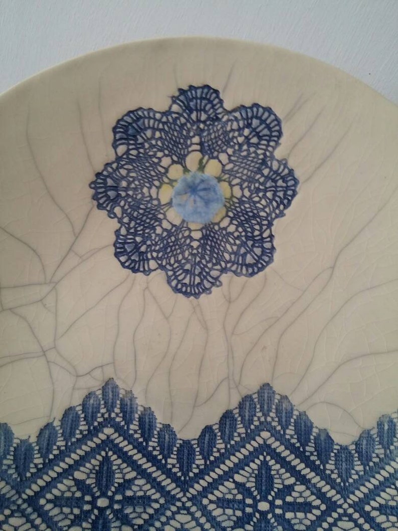 K&ouml;nnte beinhalten: Ein wei&szlig;er Keramikteller mit einem blauen Spitzenmuster. Die Spitze hat die Form einer Blume mit einer blauen Mitte. Der Teller hat eine craquel&eacute;e Glasur.