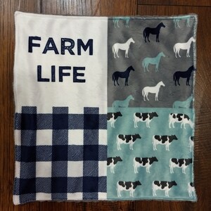 May include: A square, soft blanket with a farm theme. The blanket is divided into four sections. One section has the words "FARM LIFE" in navy blue. Another section has a navy blue and white checkered pattern. The other two sections have horse and cow patterns.