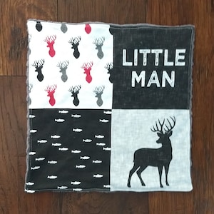 May include: A black and white baby blanket with a patchwork design. The blanket features a repeating pattern of deer heads, a row of fish, and the words "LITTLE MAN".