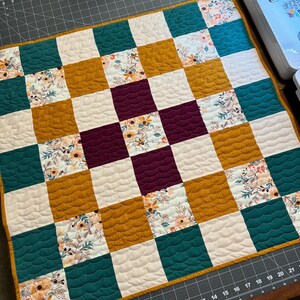 Peut inclure: Une courtepointe patchwork faite à la main avec un motif en damier. La courtepointe présente des carrés de tissus sarcelle, jaune moutarde, à imprimé floral, blanc et bordeaux. La courtepointe est bordée d'une bordure jaune moutarde.
