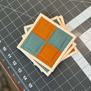 以下が含まれることがあります： 4つの正方形のキルトコースターのスタック。各コースターは、ターコイズとオレンジの正方形の2色デザインで、クリーム色のトリムで縁取られています。コースターは、グリッド線のある濃い灰色の表面に積み重ねられています。