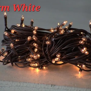 May include: A bundle of warm white string lights with small, round bulbs. The lights are on a dark brown wire and are illuminated. The words "Warm White" are in red text at the top of the image.