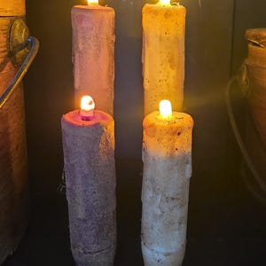 May include: Four cylindrical LED candles with a textured, weathered appearance. Two candles are a muted purple hue, while the other two are a warm cream color. Each candle features a small, glowing yellow light at the top, mimicking a flame.