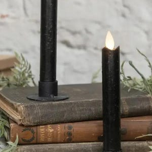 May include: Two black taper candles with textured surfaces and faux LED flames, each set in a simple black metal candle holder. The candles are positioned on a stack of three weathered, vintage-style hardcover books with visible spines.