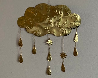 May include: A brass-colored cloud-shaped wall hanging with decorative details. The cloud has hanging teardrop and star-shaped charms suspended by chains. The wall decor is suspended by a thin, white string.