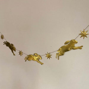 Könnte beinhalten: Eine dekorative Girlande mit goldfarbenen Hasen und sternförmigen Ornamenten, die an einem dünnen, silberfarbenen Draht aufgereiht sind. Die Hasen und Sterne sind gleichmäßig verteilt und schaffen ein skurriles und festliches Design. Ideal für Ostern oder andere Feiern.