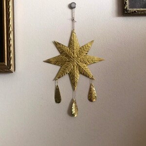 May include: A gold-colored, star-shaped wall hanging with eight points and leaf-like details. Three teardrop-shaped ornaments dangle from the star. The star is hanging on a white wall, with framed pictures in the background.