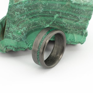 以下が含まれることがあります： 緑色のマラカイトのインレイが施された黒いカーボンファイバー製のリング。リングはマラカイトの岩の上に置かれています。