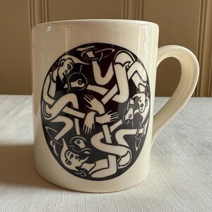 May include: A cream-colored ceramic mug with a black and white circular design featuring intertwined figures. The mug has a curved handle and is set on a white tablecloth. A potted succulent plant is visible in the background.