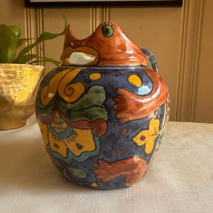 May include: A ceramic jar with a frog-shaped lid. The jar is painted with a dark blue background and colorful floral and geometric patterns in yellow, green, and orange. The lid is orange with green eyes.