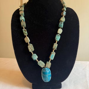May include: A necklace featuring a series of rectangular, weathered turquoise-colored beads with carved details, interspersed with small gold-toned spacer beads. The centerpiece is a larger, smooth, bright blue oval scarab beetle pendant.