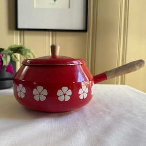 May include: A red enamel saucepan with a wooden handle and lid. The pot features white floral designs around the base. The handle and lid knob are made of wood. A decorative kitchen item.