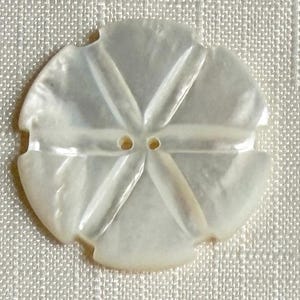 May include: A circular white mother-of-pearl button featuring a carved flower-like design with six distinct petals radiating from the center. The button has two small holes in the middle for attachment and a smooth, iridescent, glossy surface finish.